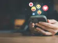 Mani che scorrono su uno smartphone, con emoji che fluttuano, simbolo di un efficace buzz marketing e interazione sui social media.