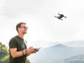 Uomo con occhiali da sole controlla un drone in volo, godendosi il panorama montano. L'utilizzo droni permette di catturare prospettive uniche.
