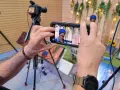Mani che tengono uno smartphone per il mobile journalism, con telecamere professionali sullo sfondo.