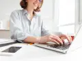 Donna sorridente al computer, che lavora per scoprire quanto si guadagna con Shopify. Sul tavolo, smartphone e documenti.