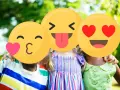 Tre bambini sorridono all'aperto, con emoji che coprono i loro volti. Un esempio di sharenting, condividere momenti felici online.