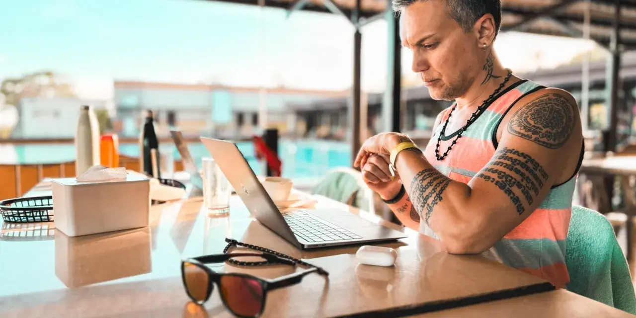 Uomo con tatuaggi lavora al laptop in un ambiente rilassato, forse un resort, mostrando la flessibilità dell'industria culturale moderna.