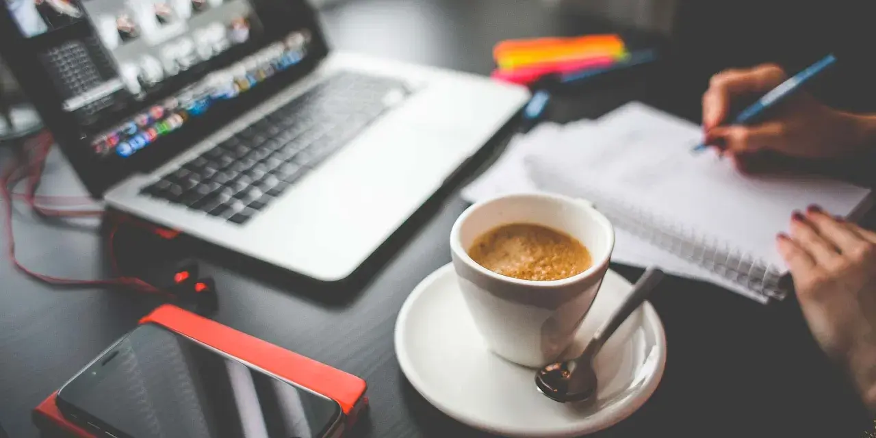 Tazza di caffè, laptop, smartphone e mani che scrivono su un quaderno. Questo è cosa significa da remoto: lavorare ovunque.