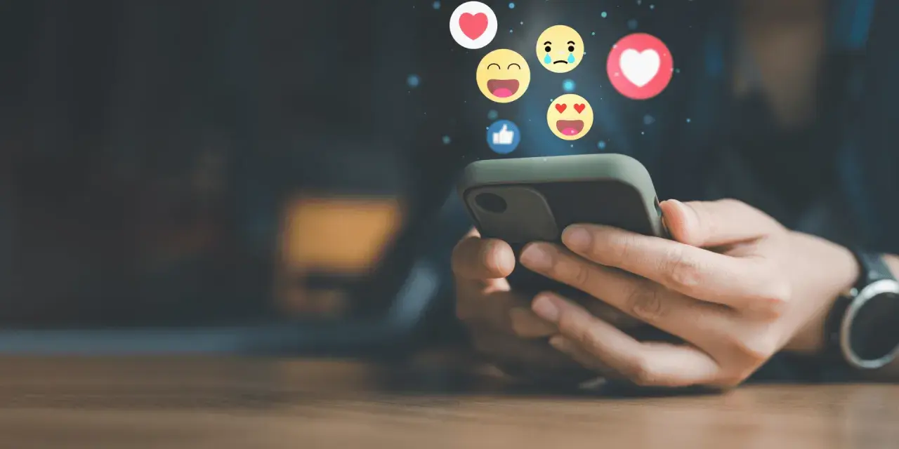 Mani che scorrono su uno smartphone, con emoji che fluttuano, simbolo di un efficace buzz marketing e interazione sui social media.