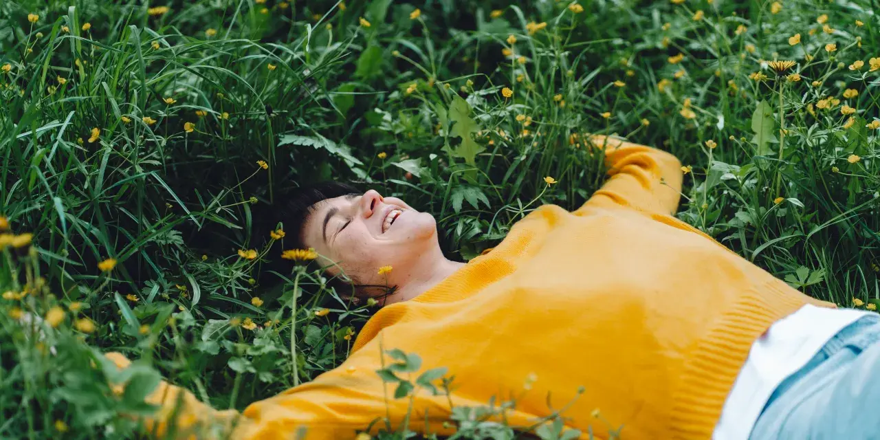 Persona sorridente sdraiata nell'erba verde con piccoli fiori gialli, godendosi un momento di digital detox.