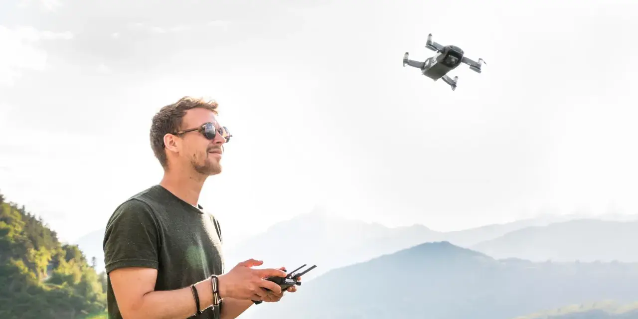 Uomo con occhiali da sole controlla un drone in volo, godendosi il panorama montano. L'utilizzo droni permette di catturare prospettive uniche.