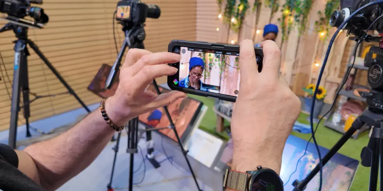 Mani che tengono uno smartphone per il mobile journalism, con telecamere professionali sullo sfondo.