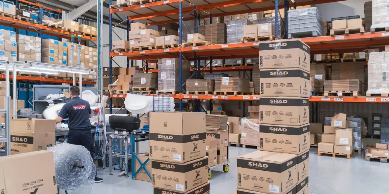 Magazzino pieno di scatoloni SHAD, pronti per spedizioni in un volatile ecommerce. Un lavoratore sistema la merce.