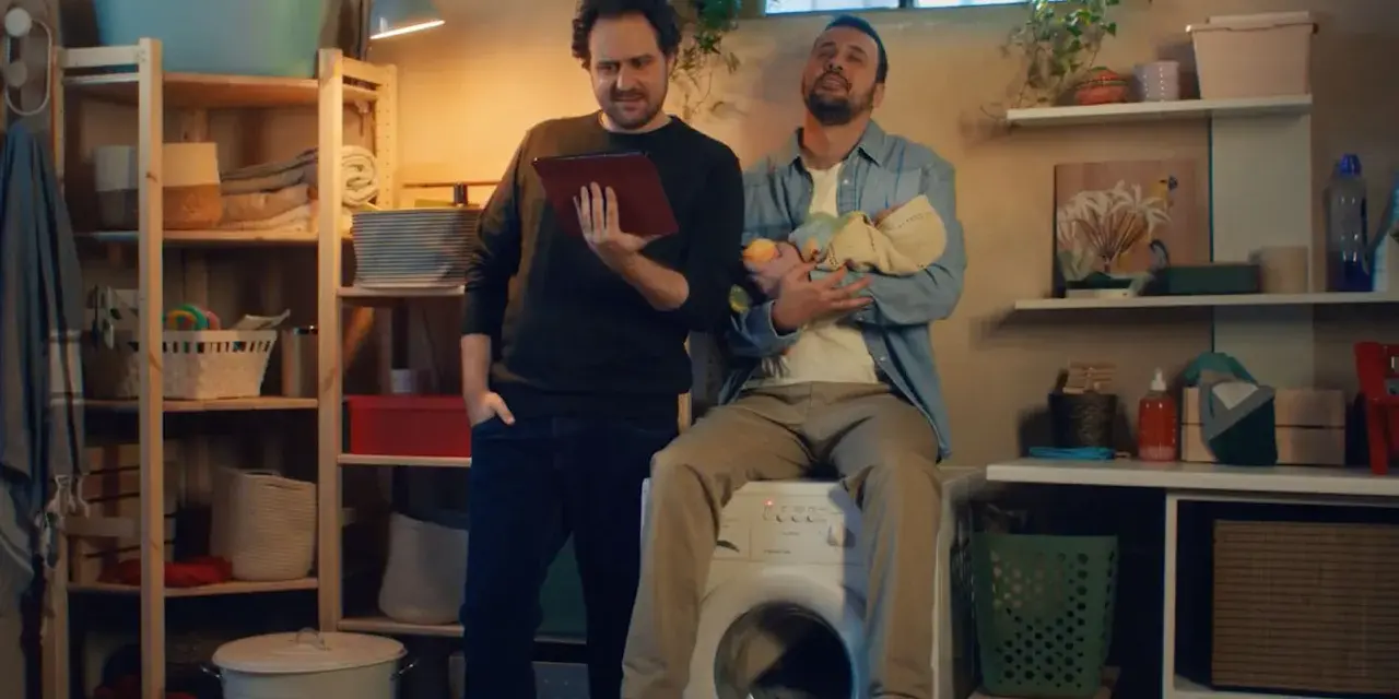 Due uomini in lavanderia, uno tiene un tablet, l'altro un neonato. Pubblicità Vodafone: la vita è più semplice con la giusta connessione.