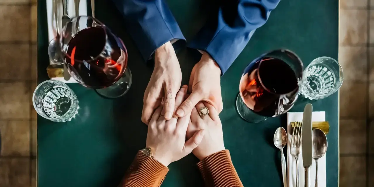 Mani che si stringono a tavola, bicchieri di vino rosso. Un momento intimo, ma il suo sguardo è perso, un red flag significato di pensieri lontani.