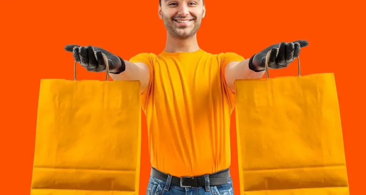 Un rider di Glovo azienda, con guanti neri e maglia gialla, porge due borse arancioni.