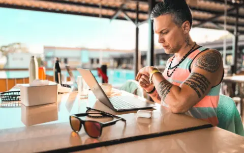 Uomo con tatuaggi lavora al laptop in un ambiente rilassato, forse un resort, mostrando la flessibilità dell'industria culturale moderna.