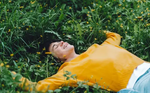 Persona sorridente sdraiata nell'erba verde con piccoli fiori gialli, godendosi un momento di digital detox.