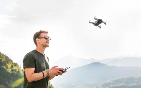Uomo con occhiali da sole controlla un drone in volo, godendosi il panorama montano. L'utilizzo droni permette di catturare prospettive uniche.