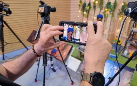 Mani che tengono uno smartphone per il mobile journalism, con telecamere professionali sullo sfondo.