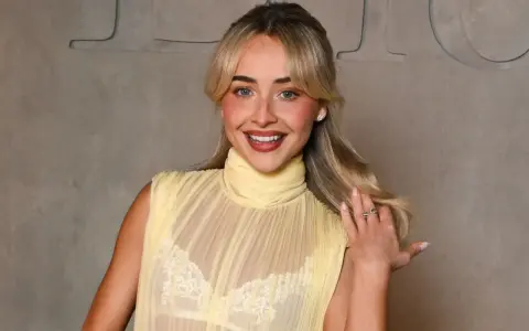 Una ragazza sorride, indossa un abito giallo trasparente con un reggiseno di pizzo bianco visibile.