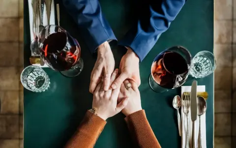 Mani che si stringono a tavola, bicchieri di vino rosso. Un momento intimo, ma il suo sguardo è perso, un red flag significato di pensieri lontani.