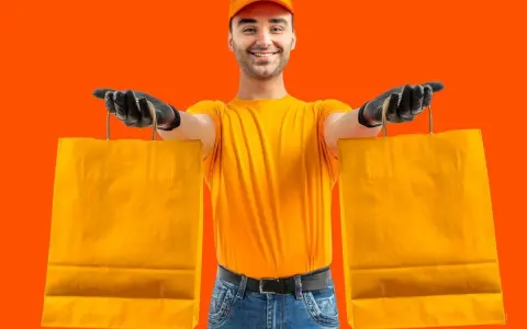 Un rider di Glovo azienda, con guanti neri e maglia gialla, porge due borse arancioni.