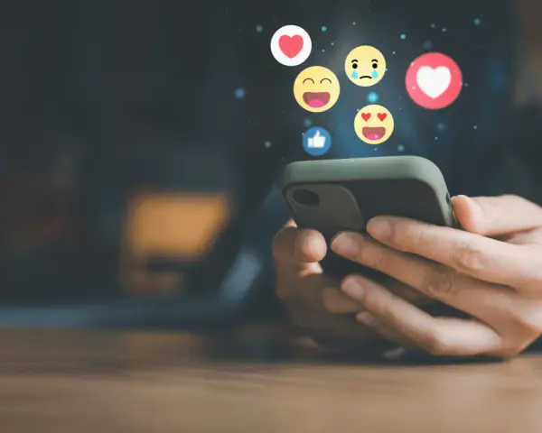 Mani che scorrono su uno smartphone, con emoji che fluttuano, simbolo di un efficace buzz marketing e interazione sui social media.