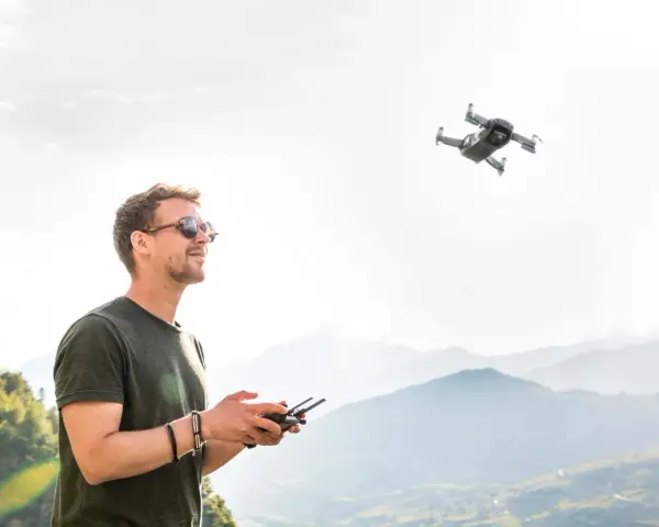 Uomo con occhiali da sole controlla un drone in volo, godendosi il panorama montano. L'utilizzo droni permette di catturare prospettive uniche.