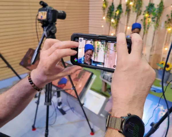 Mani che tengono uno smartphone per il mobile journalism, con telecamere professionali sullo sfondo.
