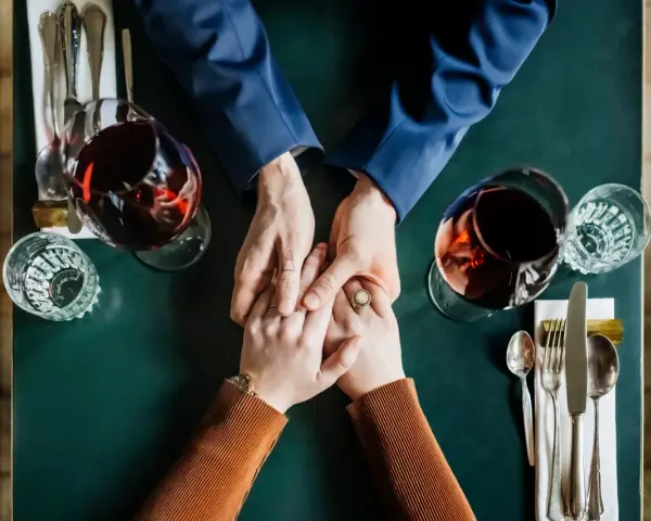 Mani che si stringono a tavola, bicchieri di vino rosso. Un momento intimo, ma il suo sguardo è perso, un red flag significato di pensieri lontani.