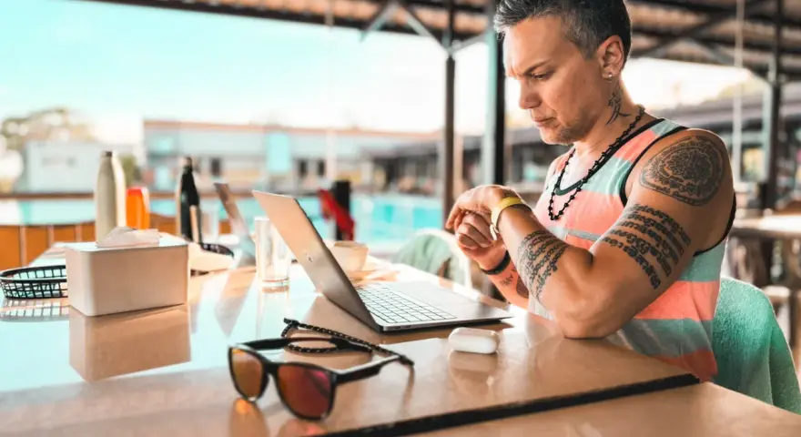 Uomo con tatuaggi lavora al laptop in un ambiente rilassato, forse un resort, mostrando la flessibilità dell'industria culturale moderna.