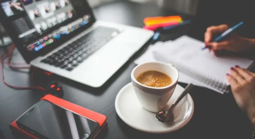Tazza di caffè, laptop, smartphone e mani che scrivono su un quaderno. Questo è cosa significa da remoto: lavorare ovunque.