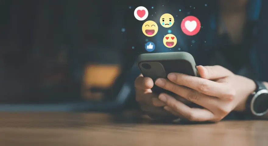 Mani che scorrono su uno smartphone, con emoji che fluttuano, simbolo di un efficace buzz marketing e interazione sui social media.