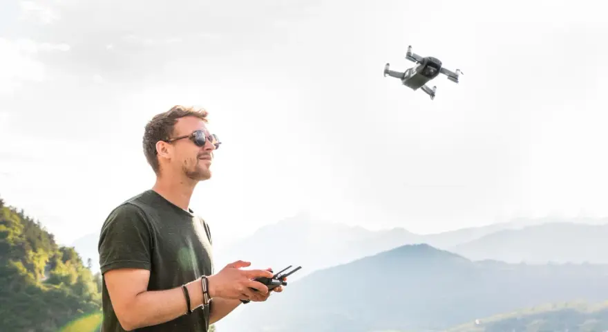 Uomo con occhiali da sole controlla un drone in volo, godendosi il panorama montano. L'utilizzo droni permette di catturare prospettive uniche.