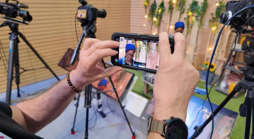 Mani che tengono uno smartphone per il mobile journalism, con telecamere professionali sullo sfondo.