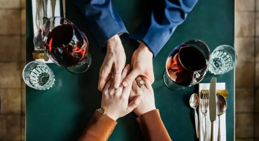 Mani che si stringono a tavola, bicchieri di vino rosso. Un momento intimo, ma il suo sguardo è perso, un red flag significato di pensieri lontani.