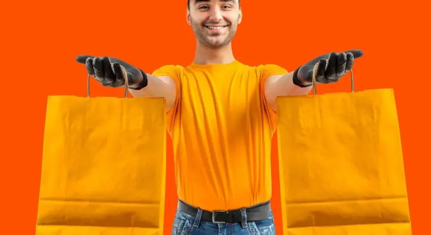 Un rider di Glovo azienda, con guanti neri e maglia gialla, porge due borse arancioni.
