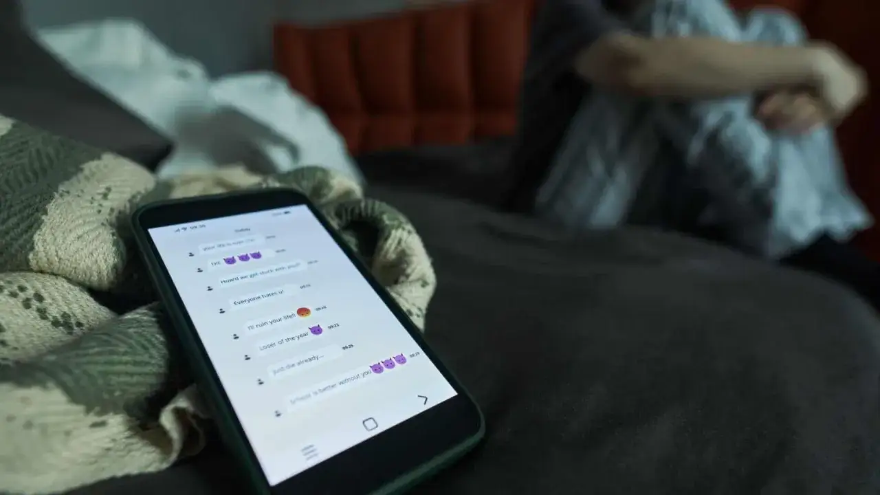 Un telefono mostra messaggi minacciosi, mentre una persona si rannicchia a letto. Un esempio di ghosting e violenza psicologica.
