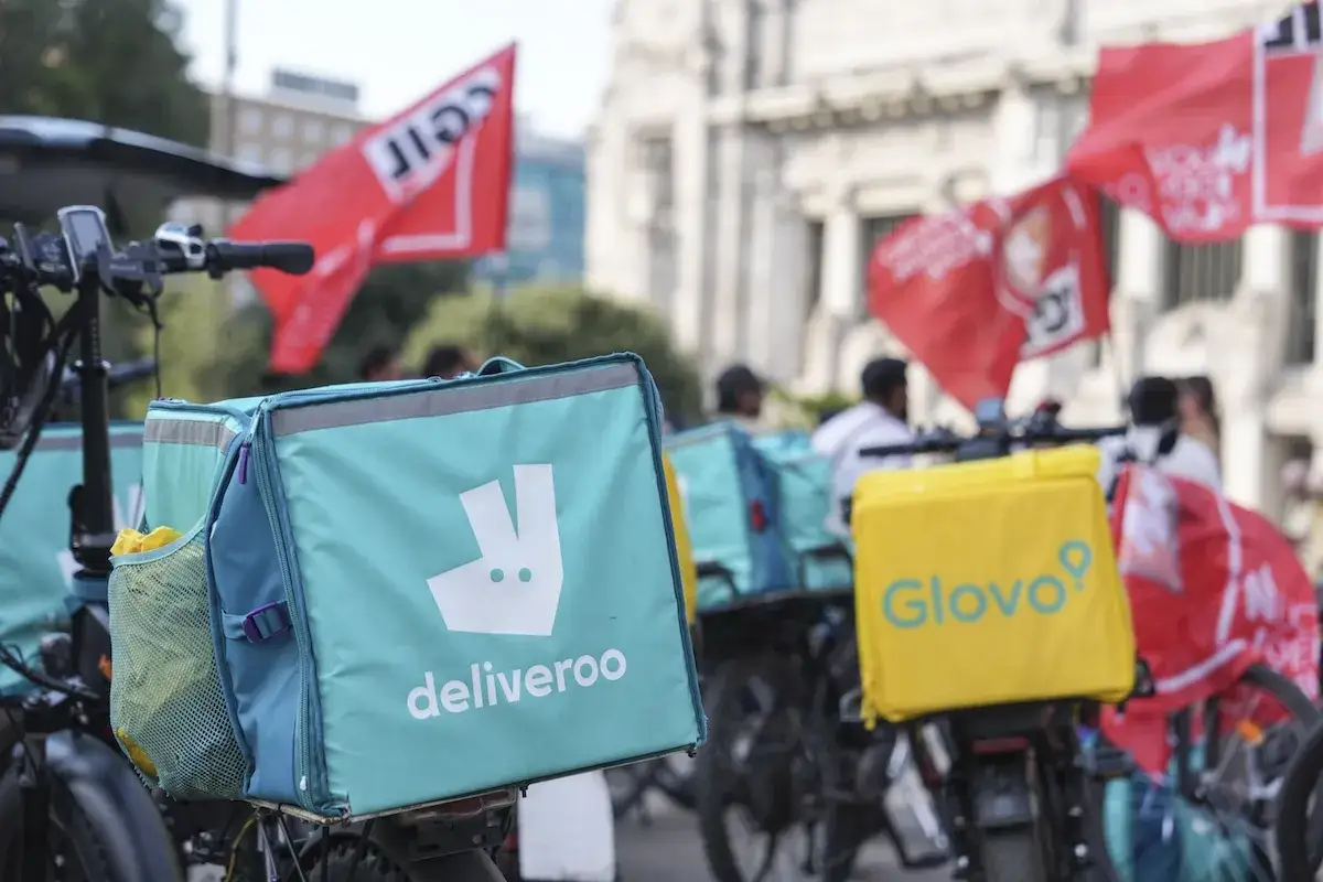 Borse da consegna di Glovo e Deliveroo in primo piano, con bandiere rosse sullo sfondo.