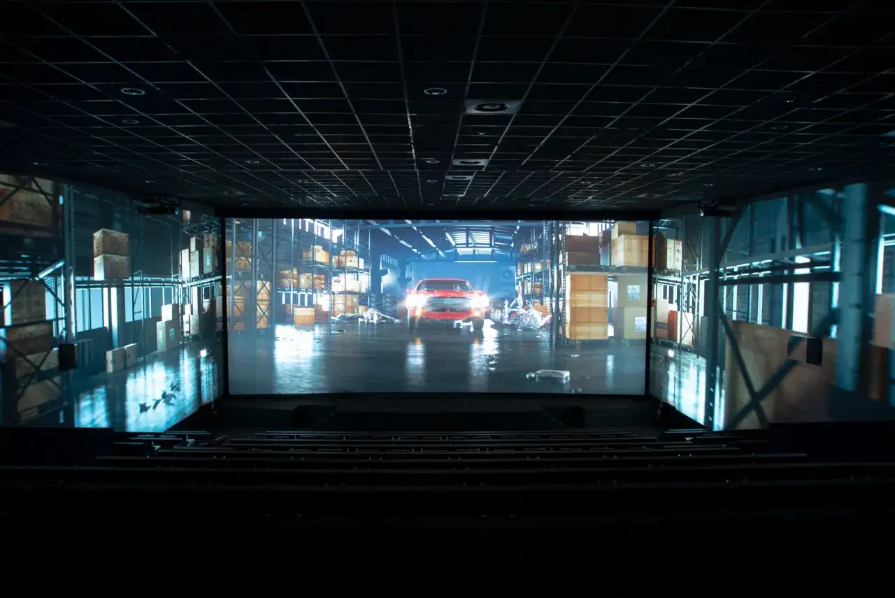 Un'auto rossa avanza in un magazzino illuminato, proiettata su un'enorme screen x cinema.