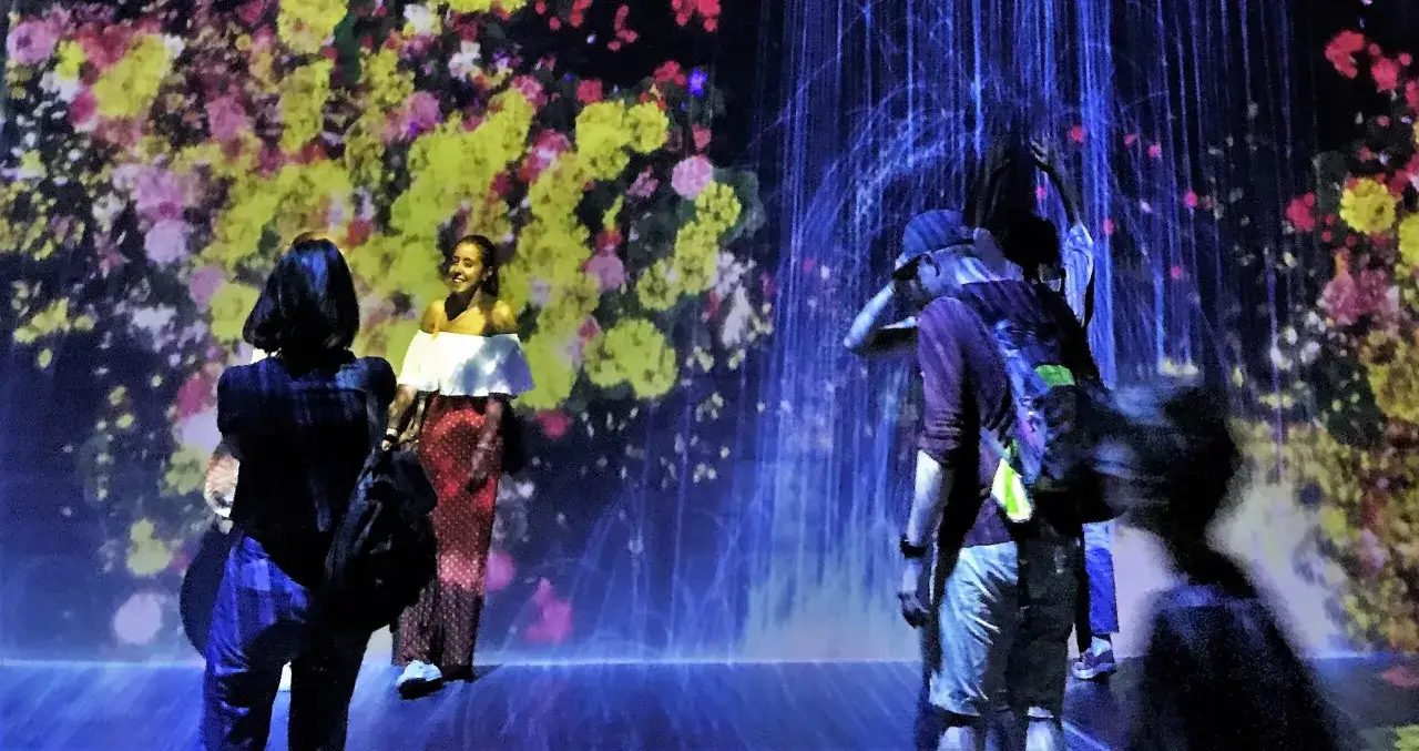 Persone ammirano un'installazione luminosa con fiori proiettati e cascate blu, un esempio di come l'industria culturale crea esperienze immersive.