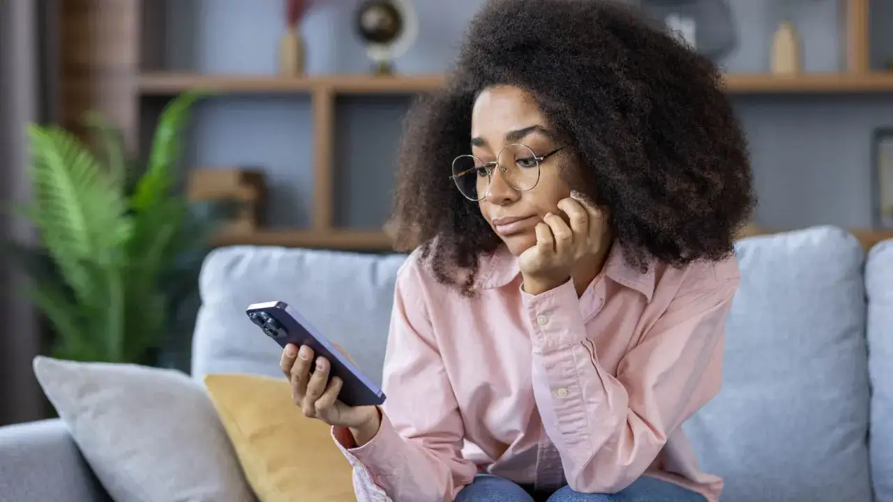 Ragazza con occhiali guarda il suo telefono, forse annoiata o pensierosa, mentre è seduta sul divano. La second screen cattura la sua attenzione.