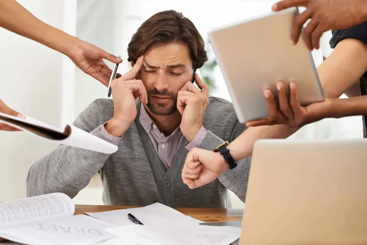 Uomo stressato da richieste multiple, simbolo dell'economia dell'attenzione. Mani che porgono tablet, fogli e penne, mentre lui si tiene la testa.